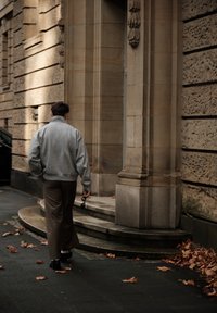 Sivá oversize mikina, hnedé nohavice s širokými nohavicami, držiace slnečné okuliare, kráčajúce po kamennom schodisku vedľa budovy s textúrovanými stenami a jesennými listami.