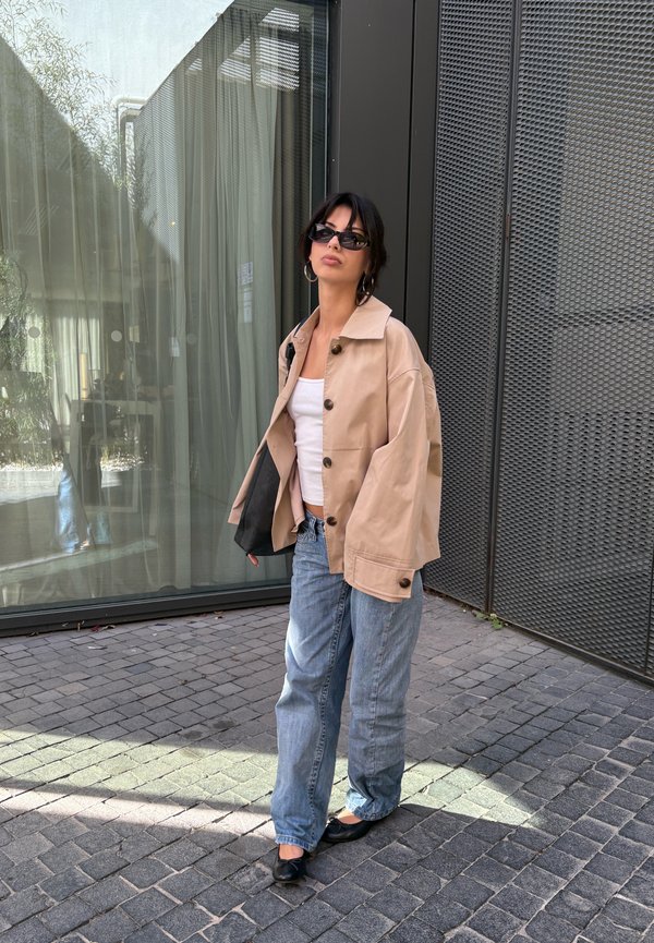 Una mujer con gafas de sol, una chaqueta beige, una camiseta blanca y vaqueros está de pie junto a un escaparate de cristal y una pared de metal oscuro.