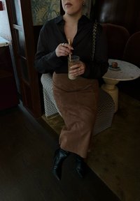 Brun semsket maxi skjørt, svart skjorte med knapper, svarte spisse støvletter. Sitter på stripete sitteplassen med et glass iskald drikke i hånden.