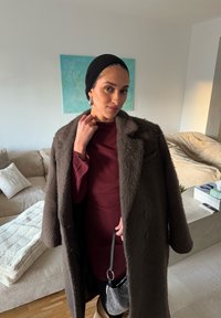 Femme à l'intérieur portant un manteau marron duveteux sur une robe bordeaux, foulard noir sur la tête, boucles d'oreilles créoles, tenant un petit sac à main noir et blanc.