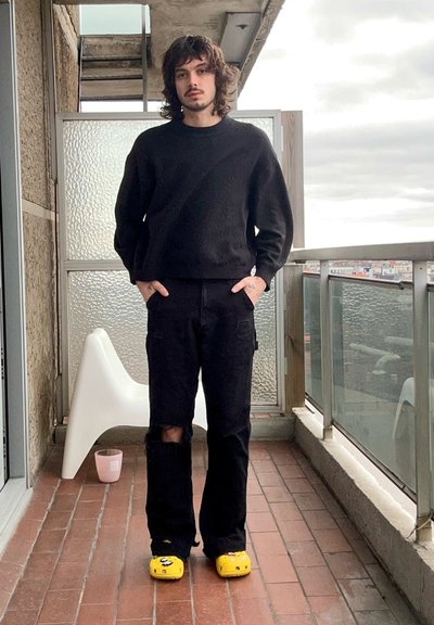 Suéter negro, pantalones cargo negros con una rodilla rasgada y zuecos amarillos con un diseño de dibujos animados. De pie en un balcón con una silla moderna.