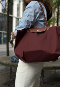 Sac fourre-tout bordeaux avec des accents en cuir marron, grande forme, texture lisse et fermeture à bouton, porté sur une chemise bleue et un pantalon blanc.