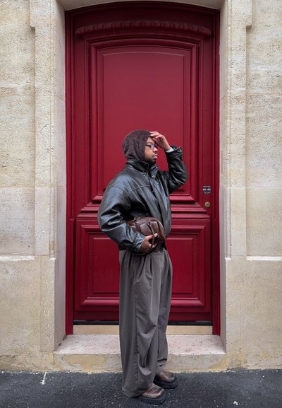 Chaqueta de cuero marrón, pantalones beige y una capucha de punto. De pie junto a una puerta de madera roja con una textura lisa y detalles sutiles en la herrajería.