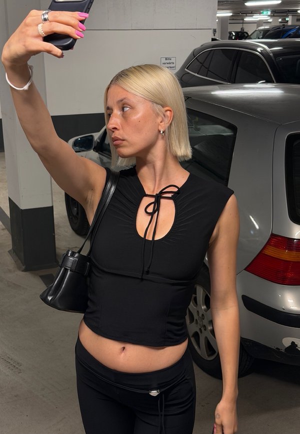 Jeune femme aux cheveux blonds courts en tenue noire prenant un selfie dans un parking faiblement éclairé à côté d'une voiture argentée.