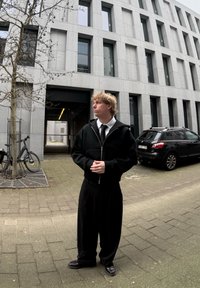 Fekete cipzáras kapucnis pulóver fehér inggel és fekete nyakkendővel, laza fekete nadrággal párosítva. Mintás járda és modern épület háttérben.