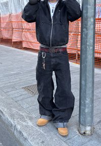 Casaco de ganga preto com fecho, grandes bolsos, combinado com calças de ganga pretas largas. Botas de trabalho castanhas e um cinto decorativo com um porta-chaves.