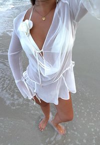 Copricostume bianco trasparente con maniche lunghe e coulisse in vita; presenta un dettaglio floreale sulla spalla. Indossato sopra un top da bagno.