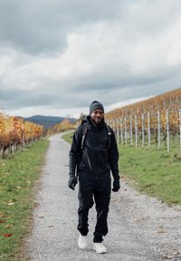 Mann mit schwarzem Beanie, Jacke, Handschuhen und Rucksack, der auf einem Kiesweg zwischen herbstlichen Weinbergen unter bewölktem Himmel spaziert.