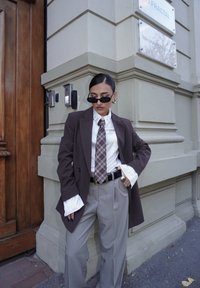 Kobieta ubrana w ciemny blazer w prążki, białą koszulę z rozkloszowanymi mankietami, kraciasty krawat, szare spodnie, okulary przeciwsłoneczne i złote kolczyki stoi przy rogu budynku.