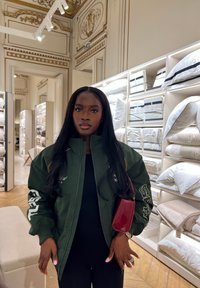 Mujer de cabello largo que lleva una chaqueta verde y sostiene un bolso rojo, de pie en una tienda de ropa de cama de lujo con estantes llenos de almohadas y sábanas.