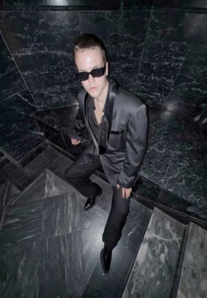 Costume noir avec des accents en satin, chemise ajustée et lunettes de soleil noires. Chaussures brillantes sur des escaliers en marbre sombre à motifs géométriques.