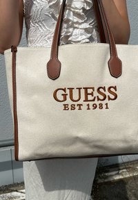Beige Baumwolltasche mit dunkelbraunen Lederakzenten. Großes "GUESS"-Logo und "EST 1981" in Braun auf der Vorderseite gestickt.
