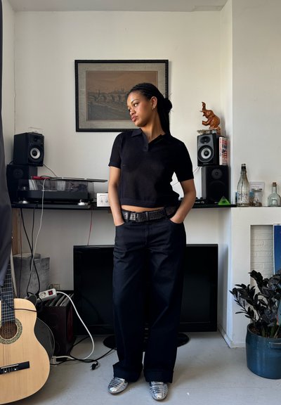 Camisa polo negra de canalé, jeans oscuros y sueltos, y zapatos plateados. El fondo cuenta con altavoces, una guitarra y una planta en maceta. Iluminación brillante.