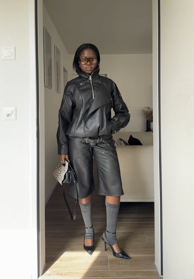 Persona con chaqueta y pantalones de cuero negro, calcetas grises hasta la rodilla, tacones negros, gafas tintadas, sosteniendo un bolso pequeño con estampado en el interior.