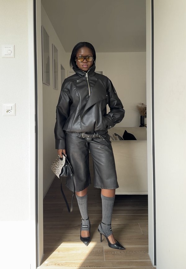 Personne portant une veste en cuir noire et un pantalon, des chaussettes grises montantes jusqu'aux genoux, des talons noirs, des lunettes teintées, tenant un petit sac à motifs à l'intérieur.