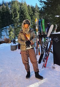 Draagt een lichtbruine, donzige jas, een grijze sjaal en bruine skipakken, staat in de sneeuw met ski's en bomen op de achtergrond.