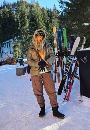 Vestido con una chaqueta acolchada marrón claro, bufanda gris y pantalones de esquí marrones, de pie en la nieve con esquís y árboles al fondo.