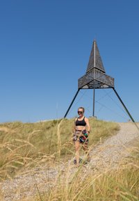 Estrutura de madeira com um telhado pontiagudo, suportes de metal preto, rodeada por relva alta. Uma pessoa caminha ao longo de um caminho de cascalho sob um céu azul claro.