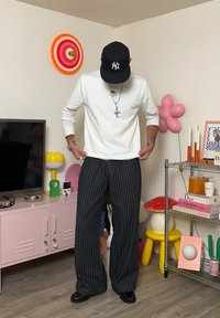 Witte long-sleeve shirt met logo, zwarte gestreepte wijde broek, zwarte pet. Achtergrond met kleurrijke decoraties en meubels.