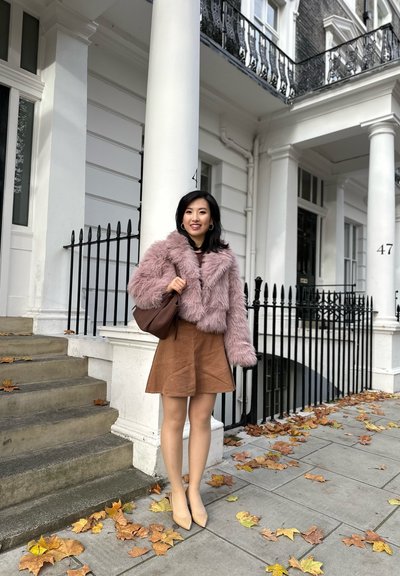 Chaqueta de piel sintética rosa, falda de pana marrón y tacones beige. Bolso marrón y hojas de otoño caídas sobre el pavimento en un entorno urbano.