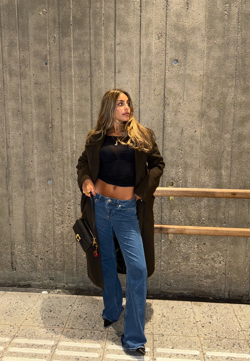 Top nero aderente, cappotto marrone oversize e jeans azzurri larghi. Gli accessori includono una piccola borsa nera con un ciondolo. Lo sfondo presenta cemento grigio.
