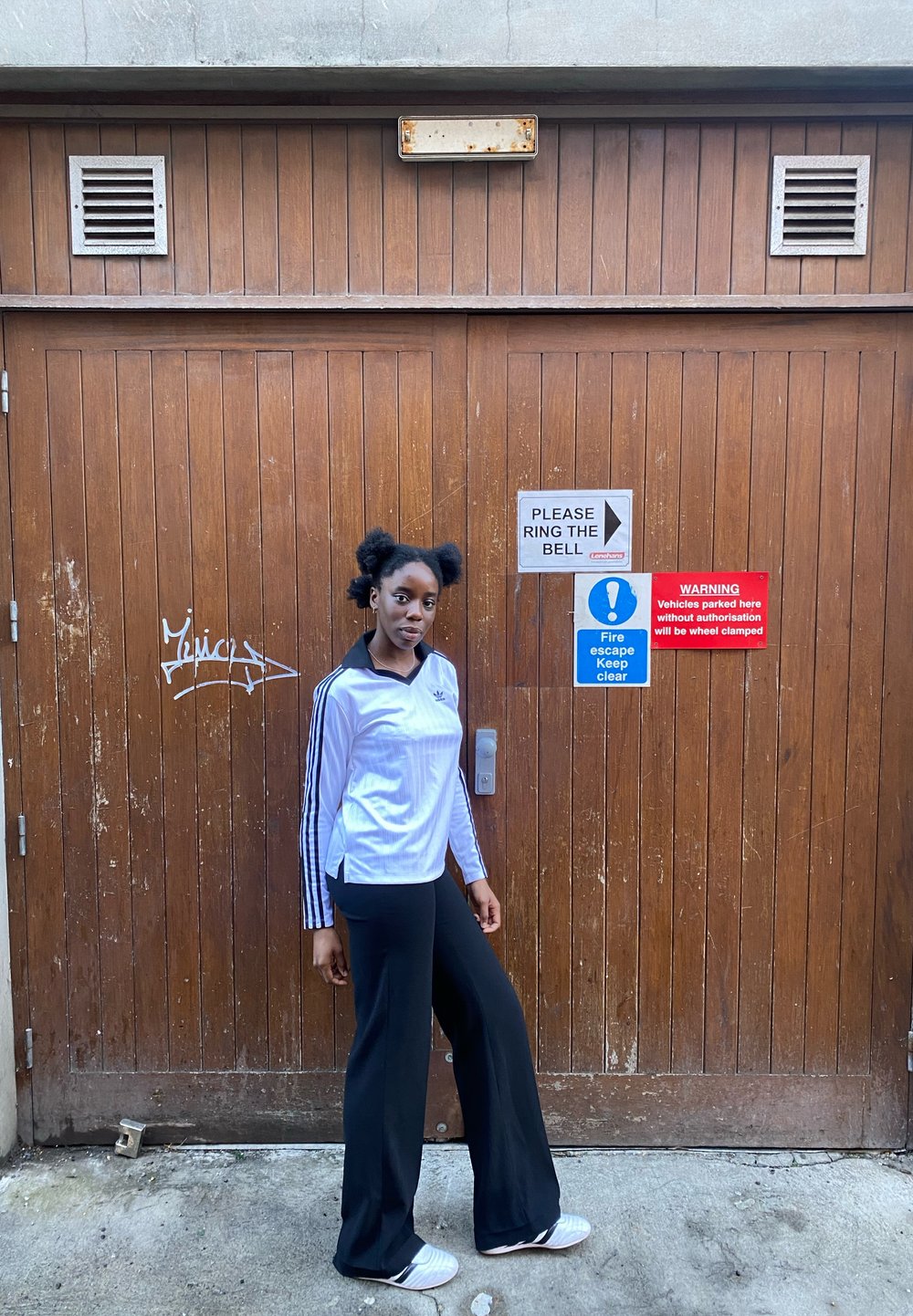 Hvid atletisk skjorte med sorte striber på ærmerne, kombineret med sorte viddebukser og hvide sko, sat op mod en trædør med skilte.