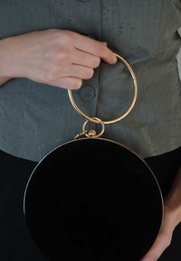 Apvali juoda rankinė su auksiniu apvaliu rankena. Pagaminta iš lygaus paviršiaus medžiagos, turi blizgų apdailą. Nešiojama su tekstūruota žalia palaidine.