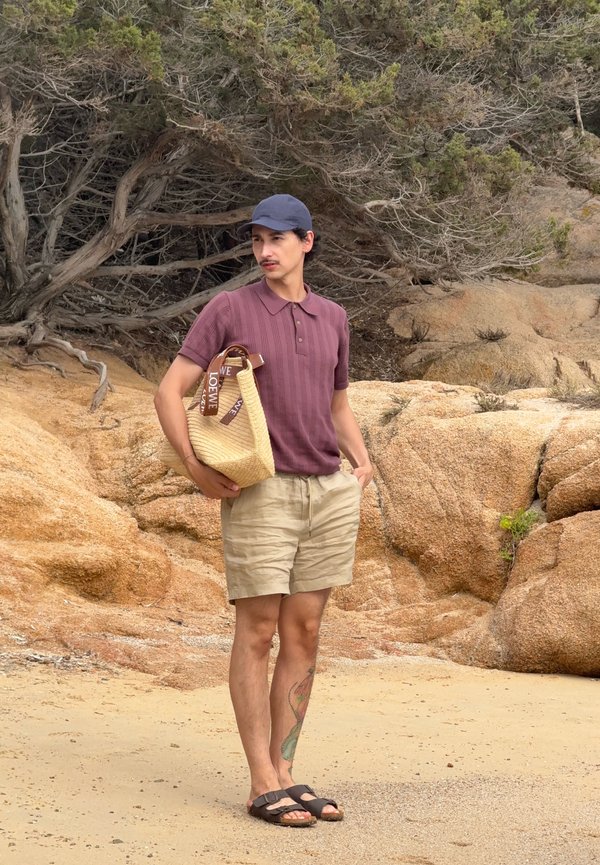 Mand iført lilla polo, beige shorts og navy kasket står på en sandstrand, mens han holder en flettet Loewe tote taske med træer og klipper i baggrunden.
