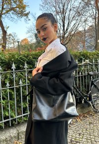 Zwarte leren tas met een gladde textuur, afgeronde vorm en minimale stiksels. Model draagt een wit shirt onder een zwarte jas.