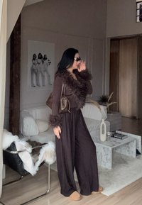 Costum căptușit maro cu accent de blană la manșete și la decolteu. Ochelarii de soare închiși la culoare și o geantă maro completează ținuta într-un interior minimalist.