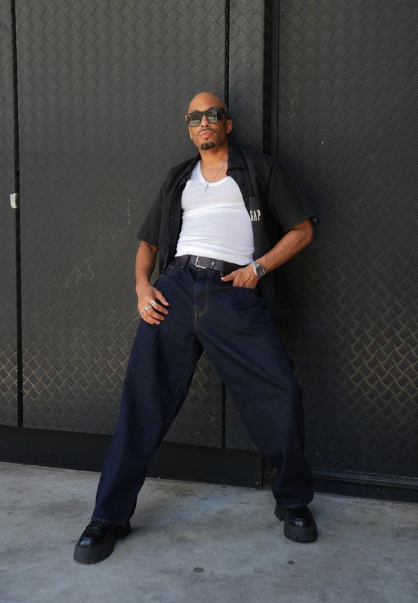 Homme portant une chemise noire ouverte sur un débardeur blanc, jeans larges foncés, chaussures noires, lunettes de soleil, debout contre un mur noir texturé, jambes écartées.