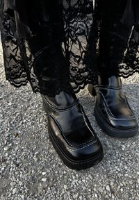 Scarpe platform nere in pelle lucida, con cuciture bianche e punta quadrata, abbinate a una gonna in pizzo nera.