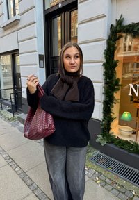 Čierny, nadýchaný sveter v kombinácii so sivými širokými nohavicami. Dr�ží veľkú, tkanú bordovú tašku. V pozadí je vyzdobené výkladné okno obchodu.