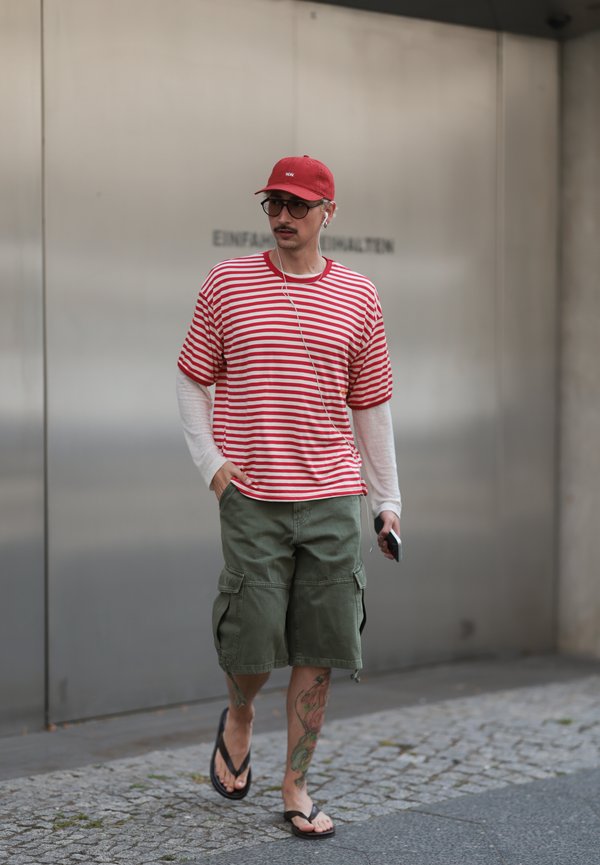 Uomo con cappellino rosso, camicia a righe rosse e bianche sopra manica lunga, pantaloncini cargo verdi, infradito e cuffie che cammina su pavimento in pietra.