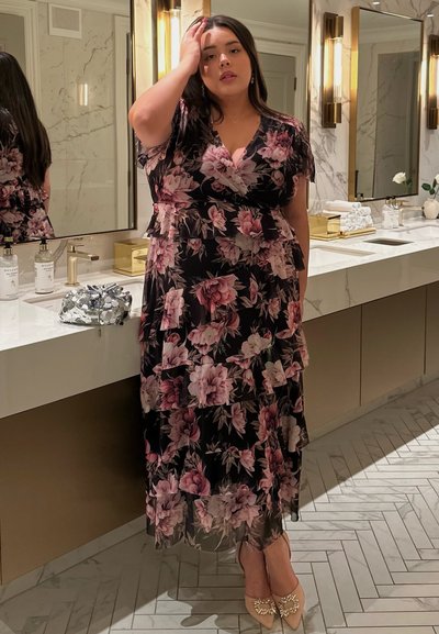 Vestido negro con flores rosas, falda con capas, mangas cortas y escote en forma de V. Zapatos beige con acentos decorativos. Escenario de baño de mármol.