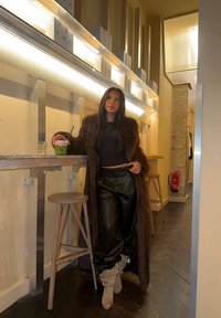 Manteau en fausse fourrure marron, pantalon large en cuir noir et bottes montantes beige. Tenant un verre vert, avec un bar en bois et des verres en arrière-plan.