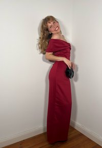 Donna in abito rosso con spalle scoperte che sorride, tiene una piccola pochette nera, in piedi contro un muro bianco su pavimento in legno.