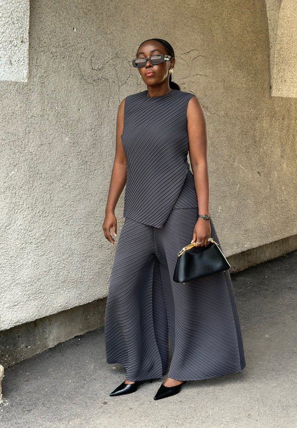 Žena oblečená v čiernom plisovanom topu a širokých nohaviciach, čierne špicaté topánky, slnečné okuliare, zlaté náušnice, drží čiernu kabelku, stojí pri textúrovanej stene.