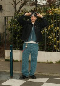 Veste en denim noire portée sur une chemise en denim bleu, associée à un pantalon bleu à motifs. Chaussures noires et chapeau pelucheux avec un logo de marque. Accessoire porte-clés.