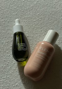 Zwei Elemis Superfood Hautpflegeflaschen auf strukturierter Oberfläche: grünes Gesichtsöl mit Pipette und pfirsichfarbige Glow-Priming-Feuchtigkeitscreme mit weißem Deckel.