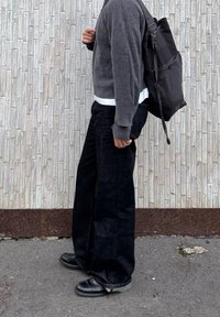 Άτομο με μαύρο παντελόνι φαρδύ, μαύρα παπούτσια, γκρι πουλόβερ και μαύρο σακίδιο, στέκεται μπροστά σε μια υφή φωτεινου γκρι τοίχου.