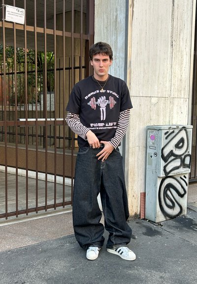 Joven con camiseta de manga larga de rayas negras y blancas en capas y camiseta gráfica negra, con vaqueros anchos oscuros y zapatillas blancas, está junto a una puerta metálica.