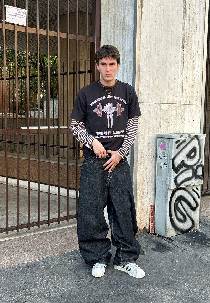Mladý muž v čiernom a bielom pruhovanom vrstvenom tričku s dlhým rukávom a čiernom grafickom tričku, široké tmavé džínsy a biele tenisky stojí pri kovovej bráne.