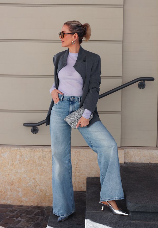 Une femme portant des lunettes de soleil pose dans un escalier. Elle porte un blazer gris anthracite, un haut clair, un jean à jambes larges et des escarpins argentés, avec une pochette à motifs.