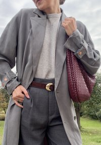 Szary, oversize'owy płaszcz trench z dużymi klapami, jasnoszary sweter, dopasowane szare spodnie, brązowy pasek oraz pleciona bordowa torebka.
