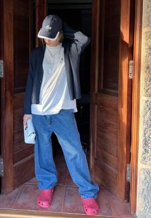 Jeans de mezclilla azul, camiseta blanca, cárdigan negro, zuecos rojos y un bolso blanco. Entorno cerca de puertas de madera con suelo de ladrillo.