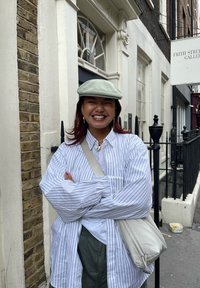 Camicia bianca a righe in taglia oversize, cappellino grigio e borsa a tracolla beige. Sorride di fronte a un muro di mattoni con un'insegna della galleria sopra.