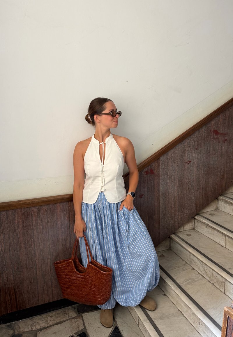 Frau trägt ein weißes ärmelloses Oberteil, einen blau gestreiften Rock und eine Sonnenbrille, hält eine braune gewebte Tasche und steht neben einer Treppe im Innenbereich.