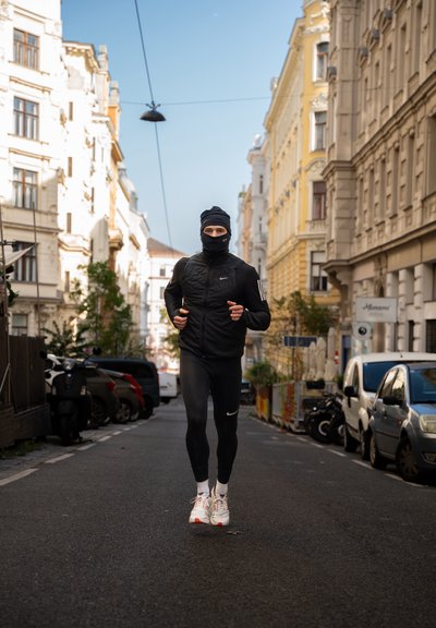 Corredor con ropa deportiva negra, que incluye una chaqueta ajustada y leggings, usando una mascarilla negra y zapatillas blancas, en una calle de la ciudad.