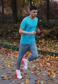 Homme athlétique courant, portant un t-shirt turquoise à manches courtes, un pantalon gris, des chaussures de course rose fluo et blanches, joggant sur un sentier couvert de feuilles.